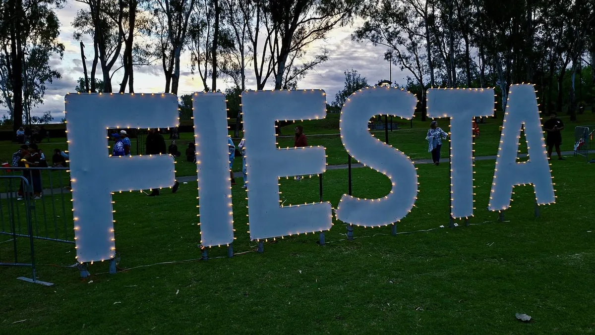 Giant letters spelling FIESTA outlined in warm lights on the grass at dusk under tall gum trees