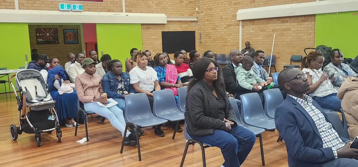 Community members seated in a school hall listening to a speaker, some holding young children